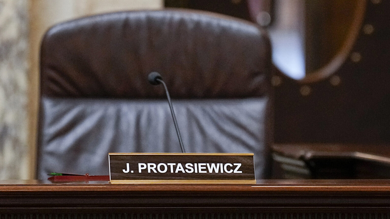 A name plate reading J. Protasiewicz sits atop the bench of a judicial dais in front of a microphone, with a high-backed leather chair and door with a vertical oval window in the background.