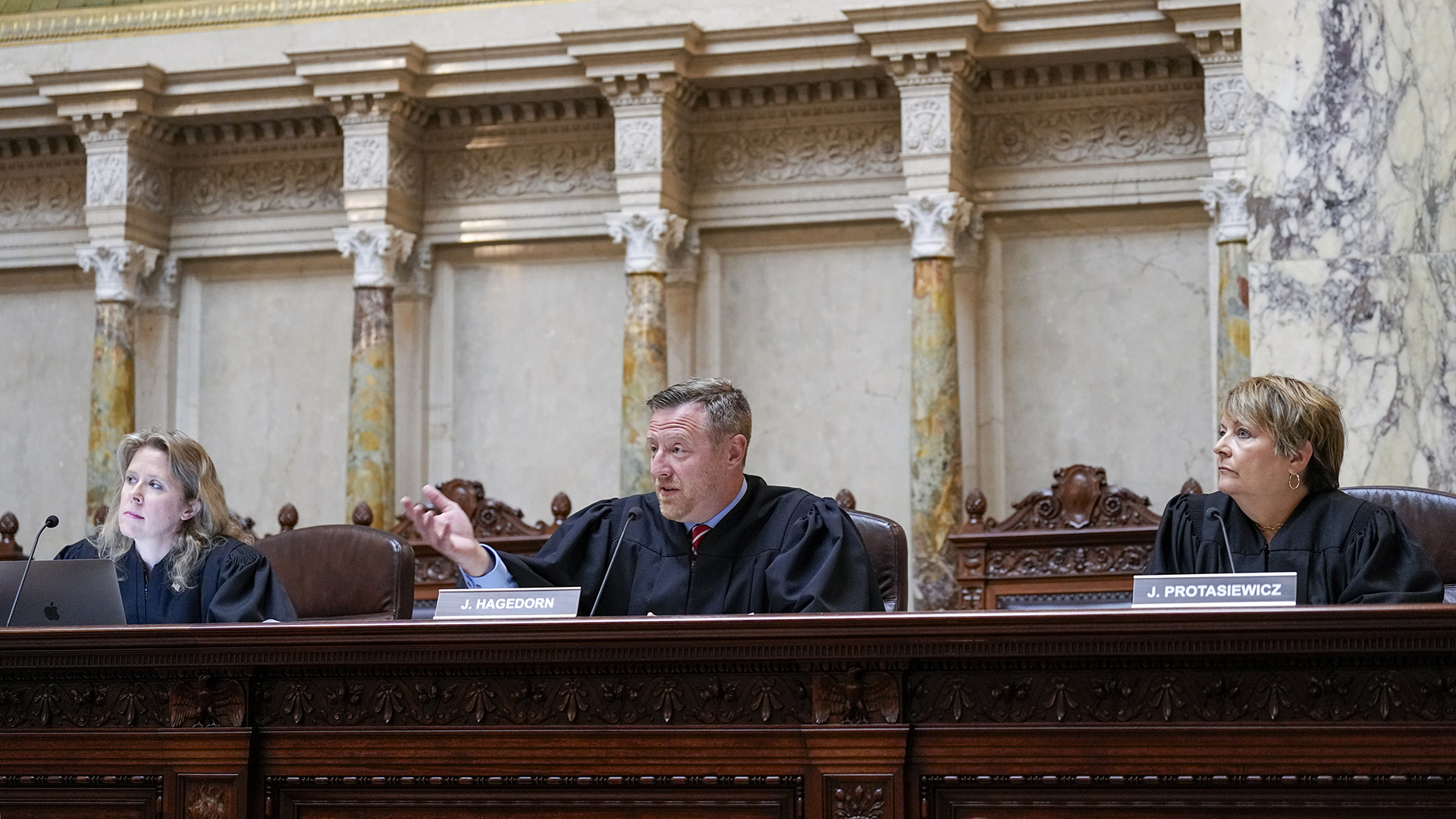 Brian Hagedorn gestures with his right hand while sitting at a judicial dais, with Rebecca Bradley and Janet Protasiewicz seated on either side, in a room with marble masonry and pillars.