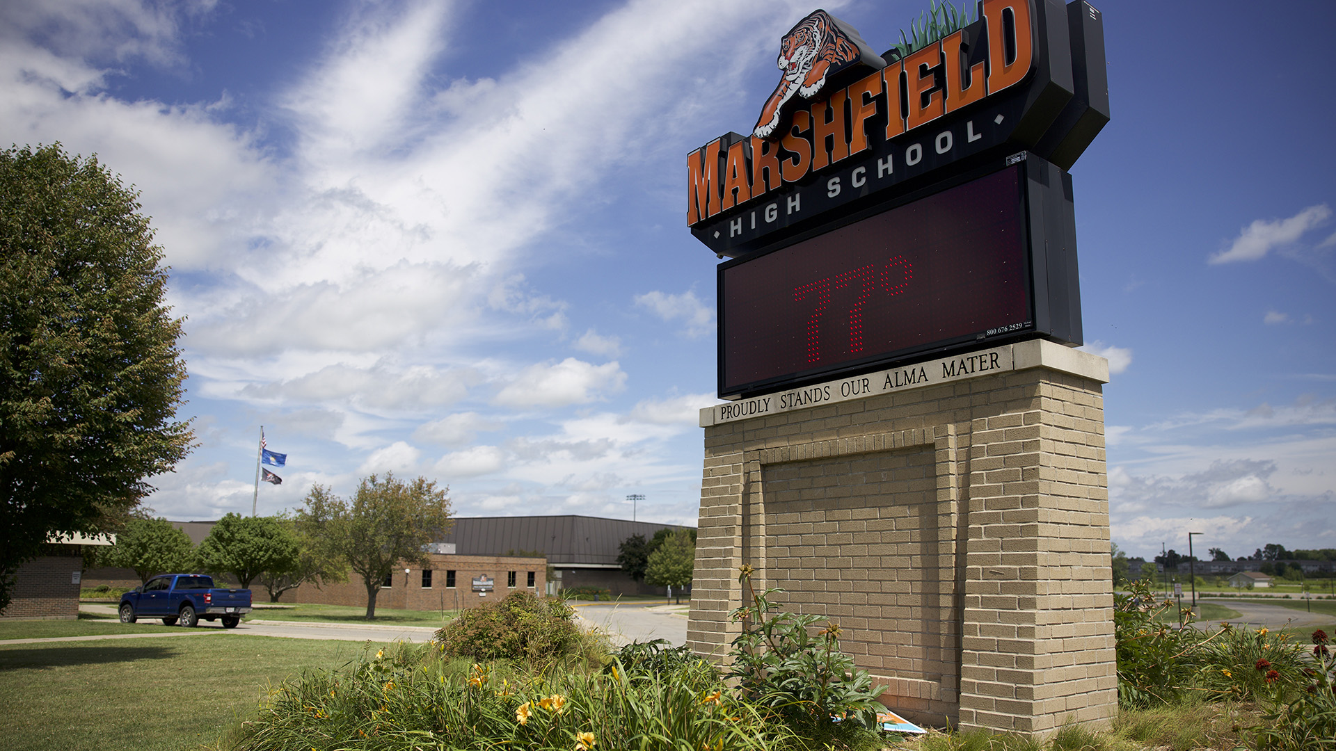 A sign with a graphic of a tiger and a wordmark reading "Marshfield High School" tops a sign with a screen showing the temperature, an inscription reading "Proudly Stands Our Alma Mater" and a brick plinth, with buildings trees and a parked vehicle in the background.