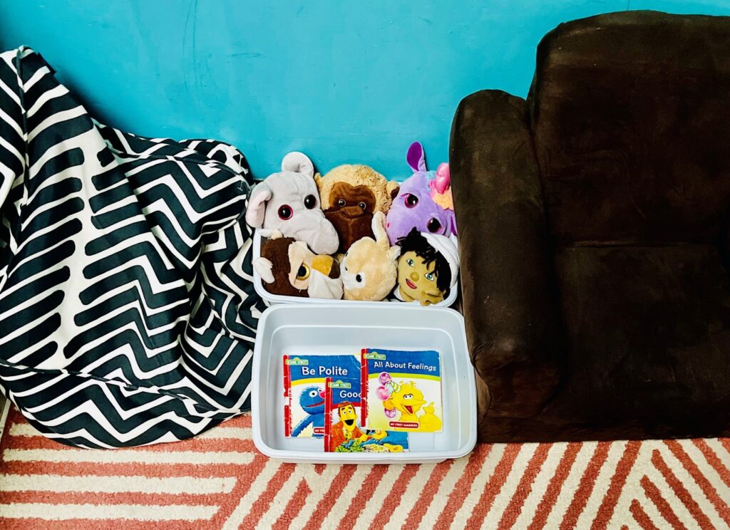 A corner of a room in a childcare center contains books, stuffed toys, and cushioned seating.