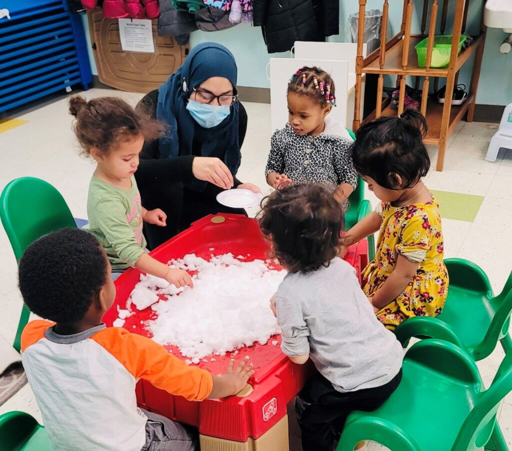 A teacher and five small children gather around a sensory table in a child care center.