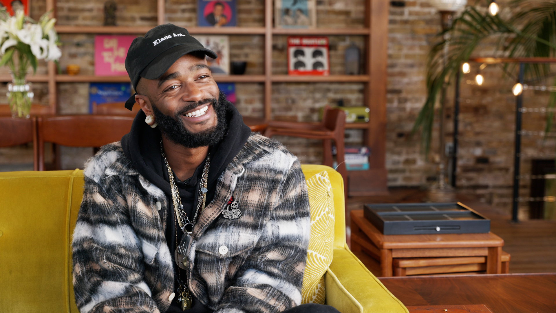 A smiling man with a beard wearing a black cap labeled 'Klass Act', a patterned fur jacket, and gold chains, seated in a cozy room with exposed brick walls, shelves displaying books and artworks, and a wooden coffee table in the foreground. The setting has a warm and welcoming ambiance with hanging lights and indoor plants.