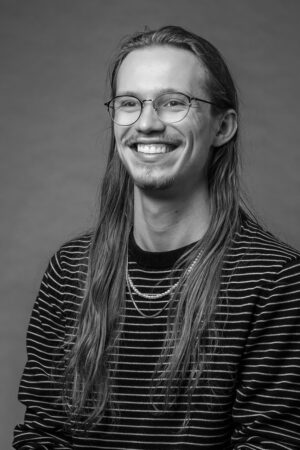 Black and white portrait of a smiling young man, Nathan Denzin, with long hair, wearing round glasses, a striped sweater, and necklaces.