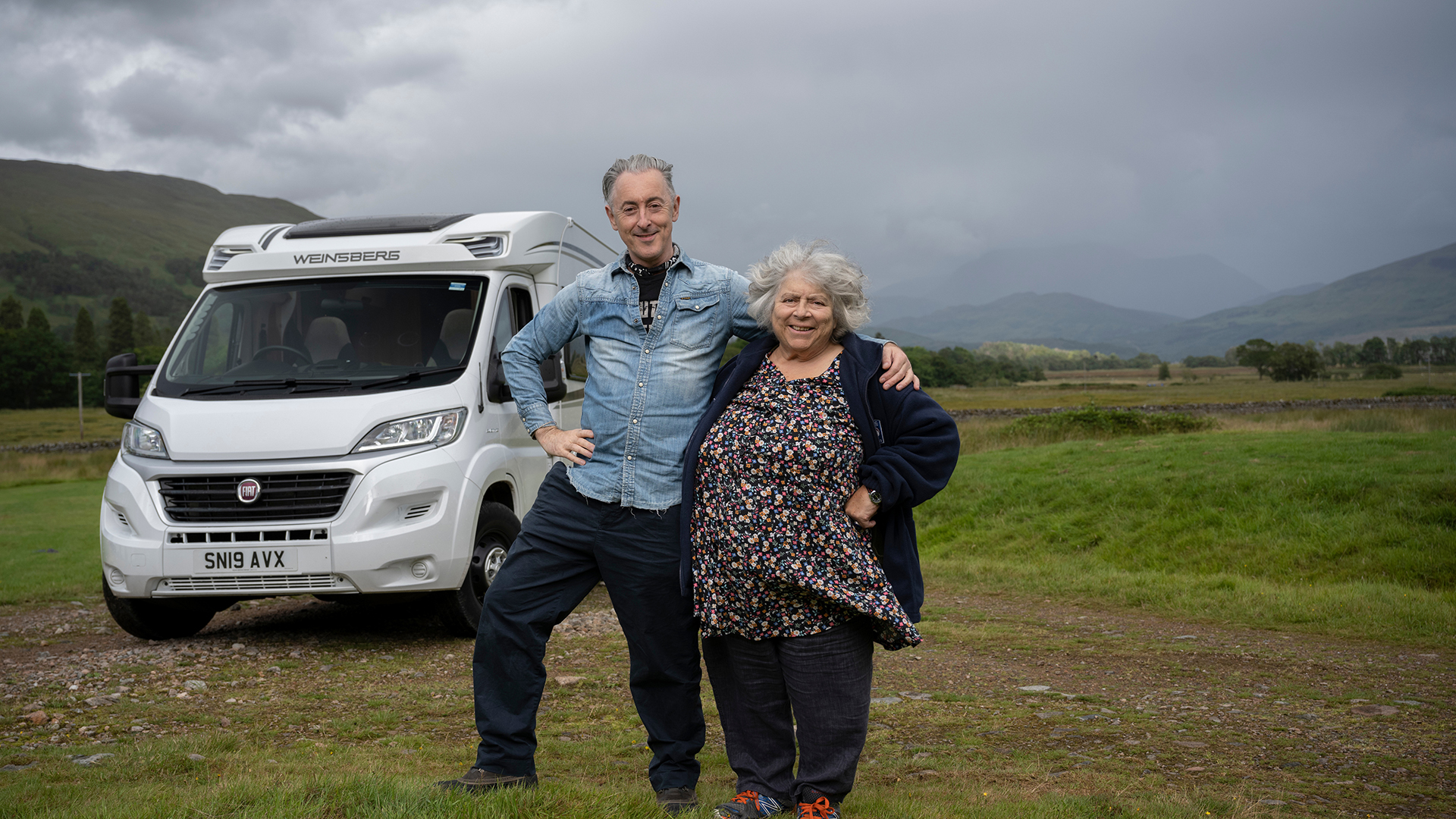 Alan Cumming and Miriam Margolyes stand with their arms around each other and smile at the camera.