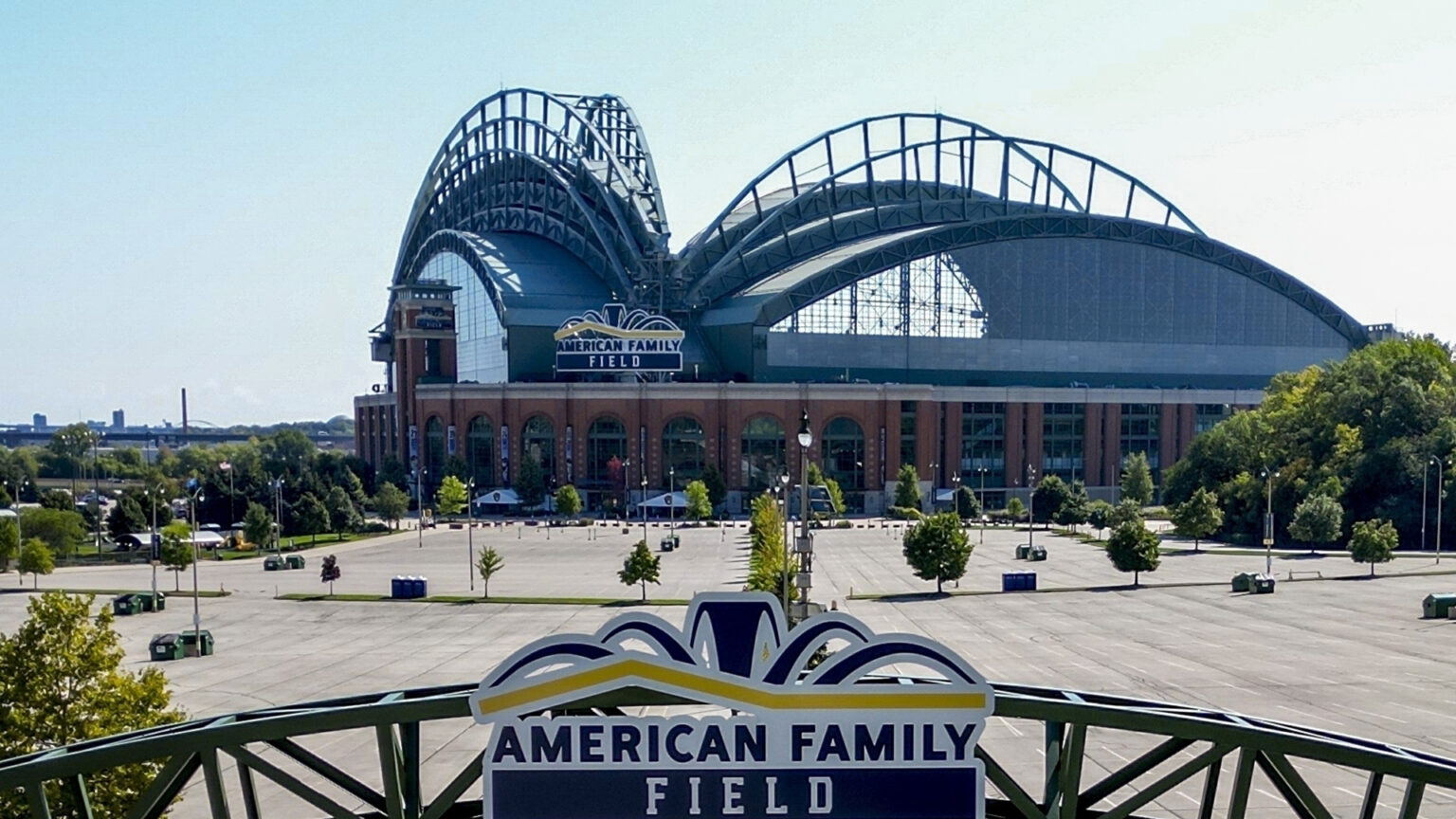 A photo shows a baseball stadium with an open retractable roof and a sign over its home plate entrance reading American Family Field, surrounded by empty parking lots and trees, with the top of a gate with another sign reading American Family Field in he foreground.