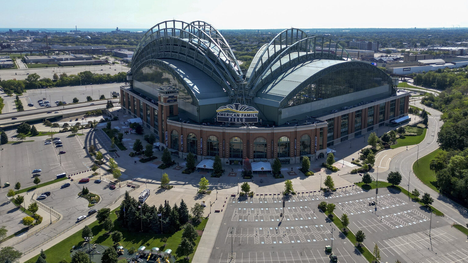 An aerial photo shows a baseball stadium with an open retractable roof and a sign over its home plate entrance reading American Family Field, surrounded by empty parking lots and roads, with an urban landscape of building and trees extending to the horizon.
