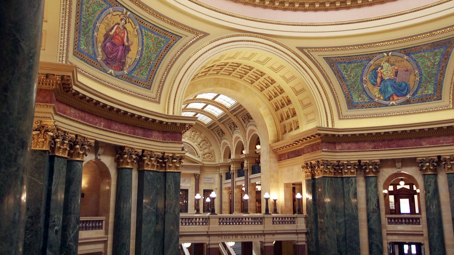 Two mosaics depicting Classical figures — one holding a scale and labeled Justice and the other holding a tablet and labeled Legislation — are seen in two quadrants of the interior walls of a rotunda inside a building with marble pillars and other masonry in a Neoclassical design.