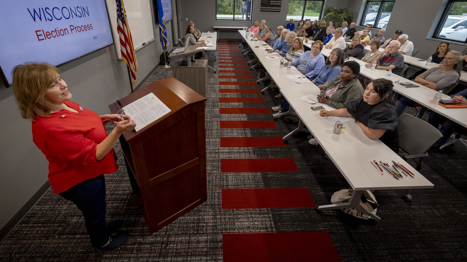 Kathy Bernier stands behind a wood podium and speaks while gesturing with both hands, facing dozens of people seated at three long tables, with several other people seated at another table facing the audience, in a room with a monitor displaying the words Wisconsin Election Process, the U.S. and Wisconsin flags, a decoration of a U.S. flag with one white stripe colored red and blue, and windows facing a parking lot filled with vehicles.