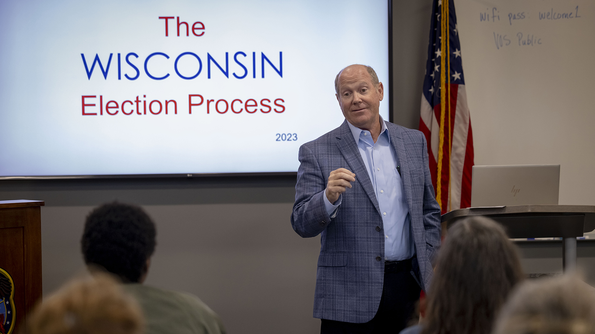 Reid Ribble stands and speaks to a group of people facing him, with the backs of several out-of-focus heads in the foreground, in a room with a wall-mounted monitor showing the words "The Wisconsin Election Process 2023," a U.S. flag, and a whiteboard.