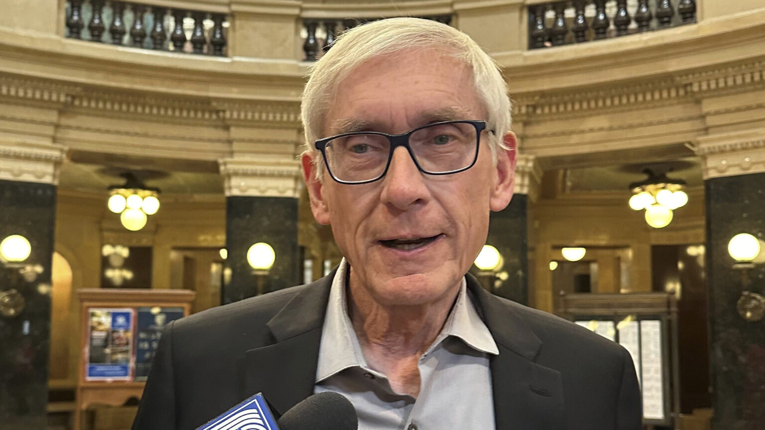 Tony Evers speaks into a hand-held microphone while standing in a room with square marble pillars framing a circular space with a marble balustrade on a second level, with numerous brass-mounted lights on the pillars, walls and ceiling.