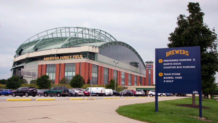 A sign labeled Brewers and with arrows pointing toward different locations stands beside a row of trees and next to a parking lot in front of a baseball stadium with a closed retractable roof and a sign reading American Family Field under a cloudy sky.