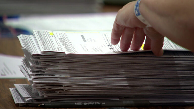 A hand pulls an absentee ballot envelope from the top of a stack of ballot envelopes on a table.