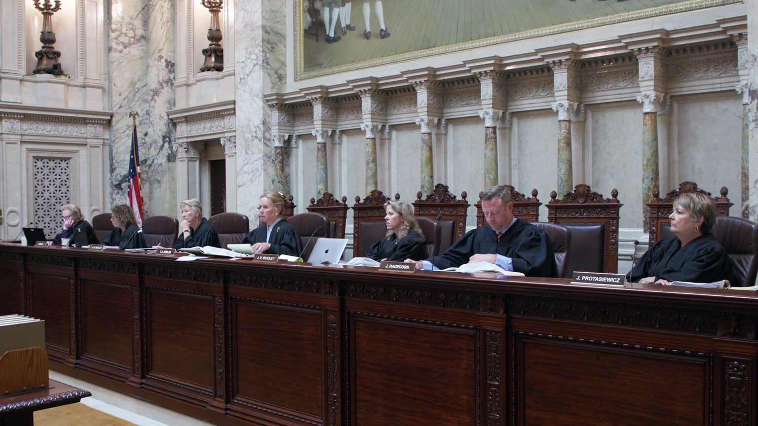 From left to right, Jill Karofsky, Rebecca Dallet, Ann Walsh Bradley Annette Ziegler, Rebecca Bradley, Brian Hagedorn and Janet Protasiewicz sit at a judicial dais in a row of high-backed leather chairs behind them, with another row of high-backed wood and leather chairs behind them, in a room with marble masonry, wall-mounted metal light fixtures, a U.S. flag and a large painting.