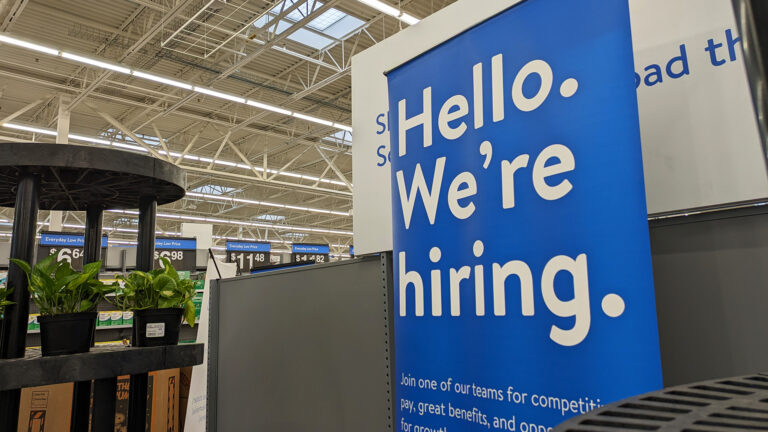 A sign with the words Hello. We're hiring. stands next to a divider in a warehouse-style store next to a stand with potted plants, with price tag signs in the background.