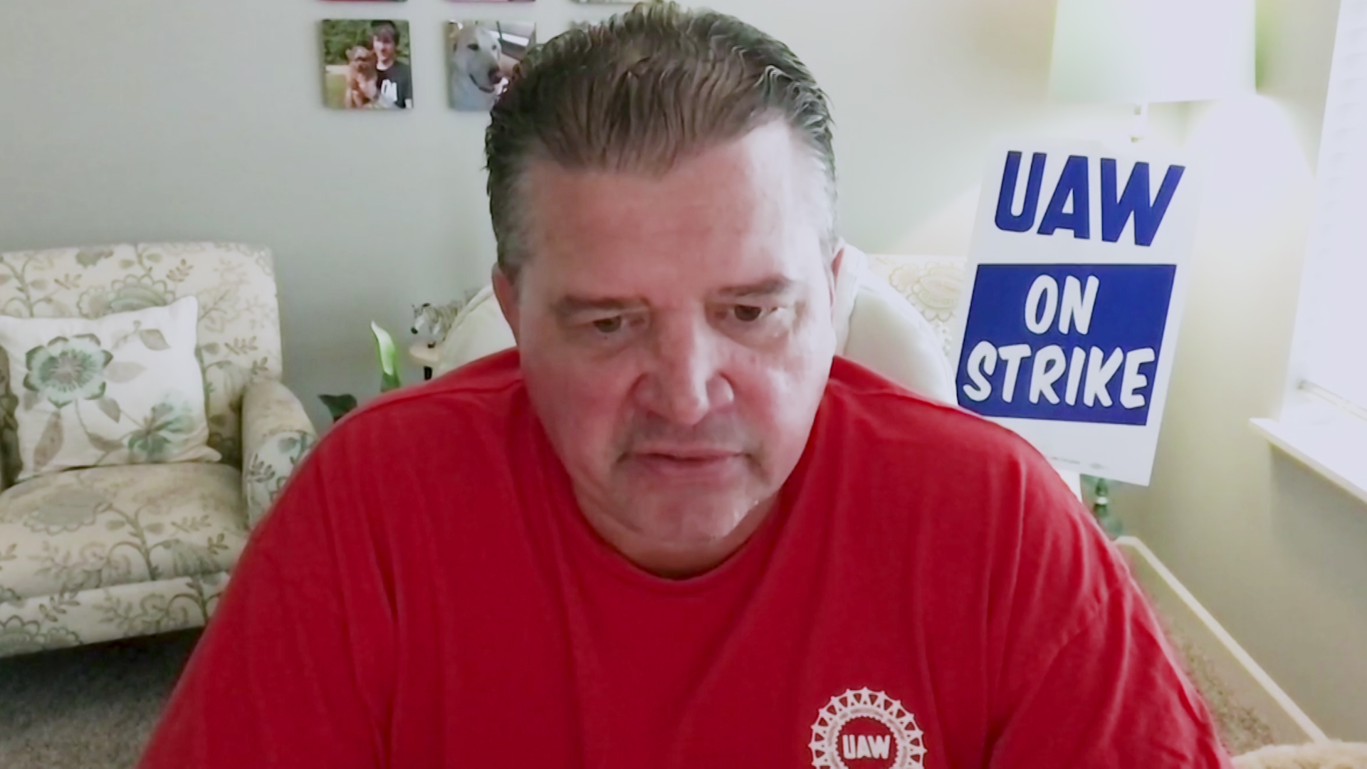 Steve Frisque speaks while facing a computer camera in a room with a lamp, furniture, photos on the wall, and a placard reading "UAW On Strike."