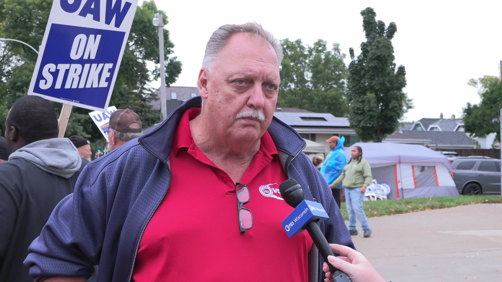 Joe Neu speaks into a microphone with a PBS Wisconsin flag while standing outside, with other people standing behind him with picket signs reading "UAW On Strike," with a tent, vehicles and houses in the background.