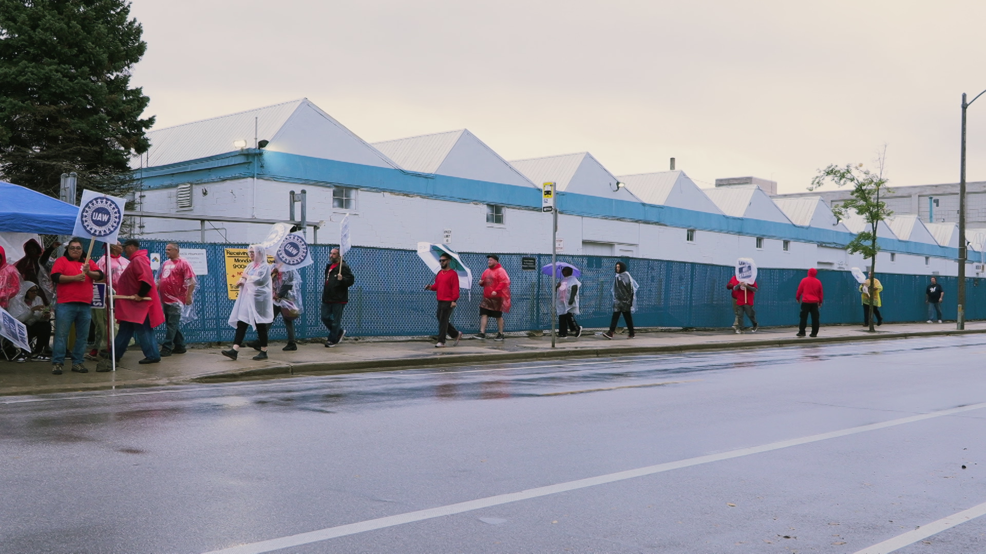 People wearing ponchos and raincoats and carrying umbrellas and signs with the United Auto Workers gear logo walk along a sidewalk, with a rain-slicked street in the foreground, and a chain-link fence with privacy slats and topped with barbed wire in front of a building with a saw-tooth roof in the background.