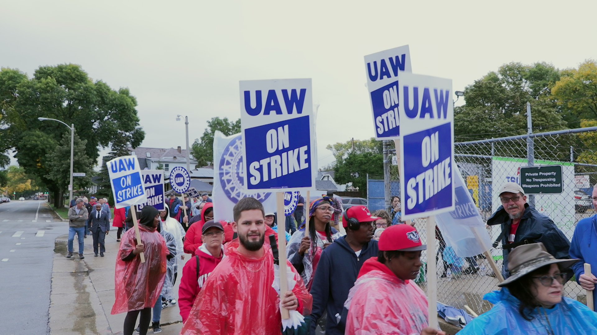 People wearing ponchos carrying signs with the United Auto Workers gear logo and the words "UAW On Strike" walk along a sidewalk next to a chain-link fence topped with barbed wire, with a street, building and trees in the background.