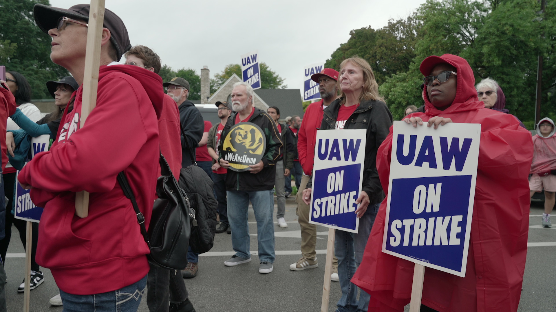 A group of people wearing ponchos and raincoats and holding signs reading "UAW On Strike" stand outside and face the same direction while listening to a speaker, with a house and trees in the background.