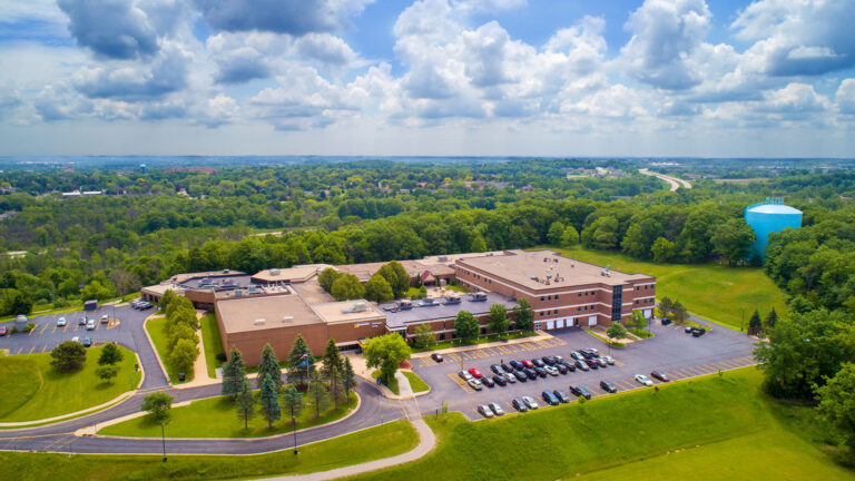 An aerial photo shows a series of connected two-and-three story brick building, surrounded by parking lots, lawns, and a wooded area with a water tower, with a divided highway, buildings and trees extending to the horizon under a partly cloudy sky.