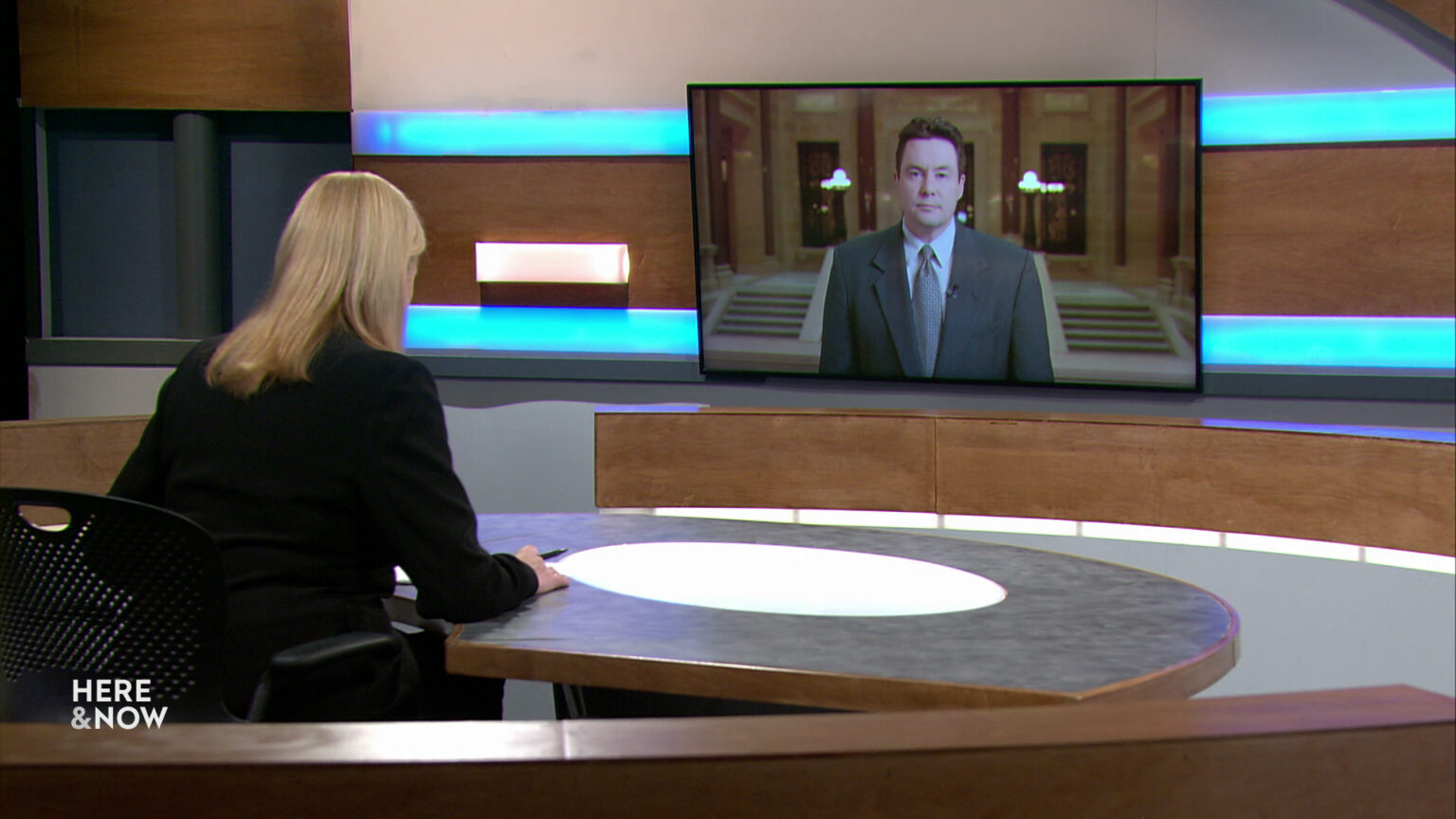 Frederica Freyberg sits at a desk on the Here & Now set and faces a video monitor showing an image of Zac Schultz.