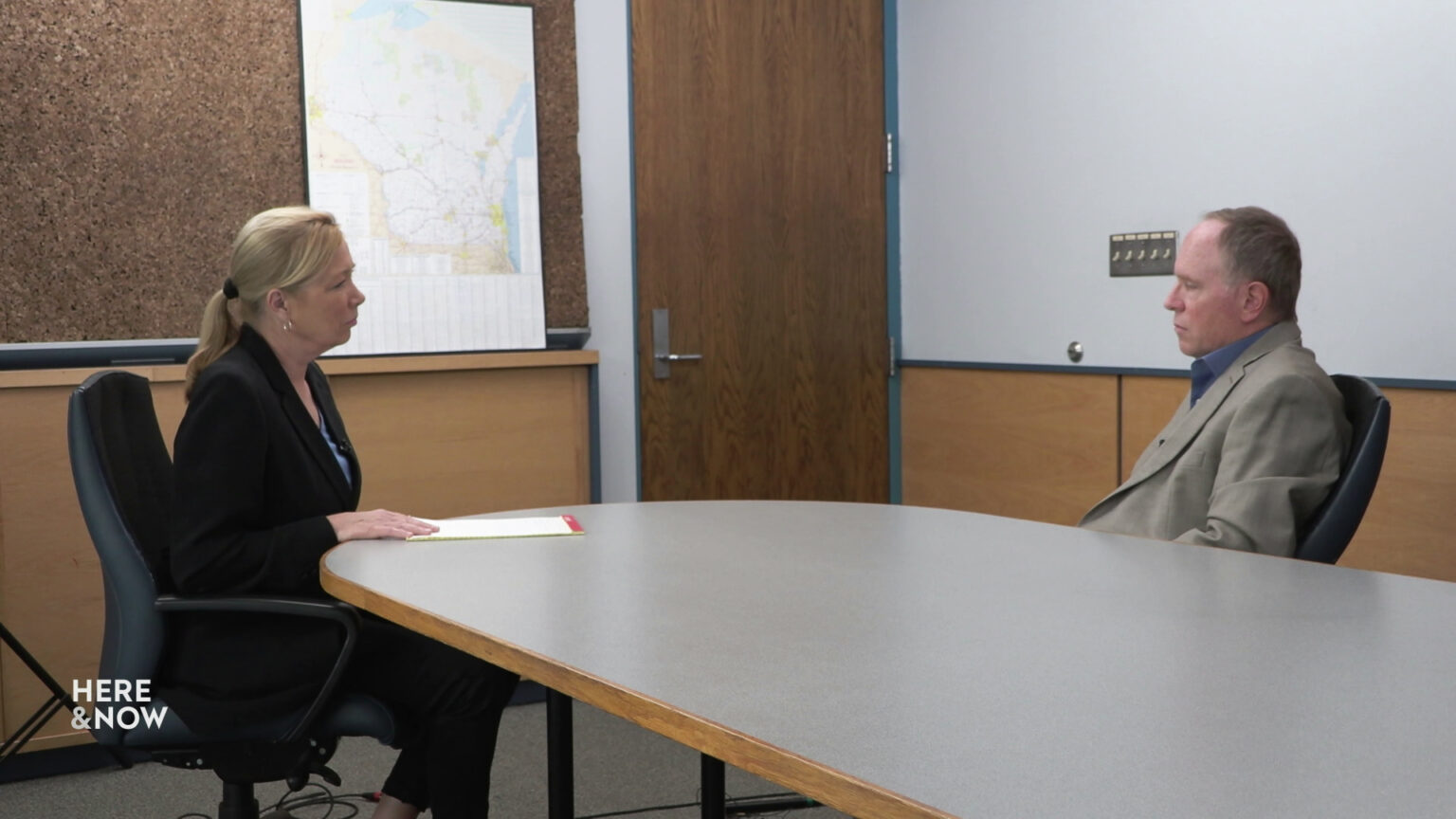 Tommy Thompson and Steve Wildeck sit facing each other across a table in a room with wood-paneled lower walls, a corkboard and map of Wisconsin, and a closed door.