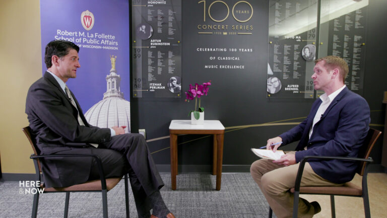 Paul Ryan and Steven Potter face each other while seated in stackable chairs in a room with a table topped by an orchid, a banner with an image of the Wisconsin State Capitol dome and the words Robert M. La Follette School of Public Affairs, and a commemorative installation on the rear wall referencing classical music excellence with a list of names.