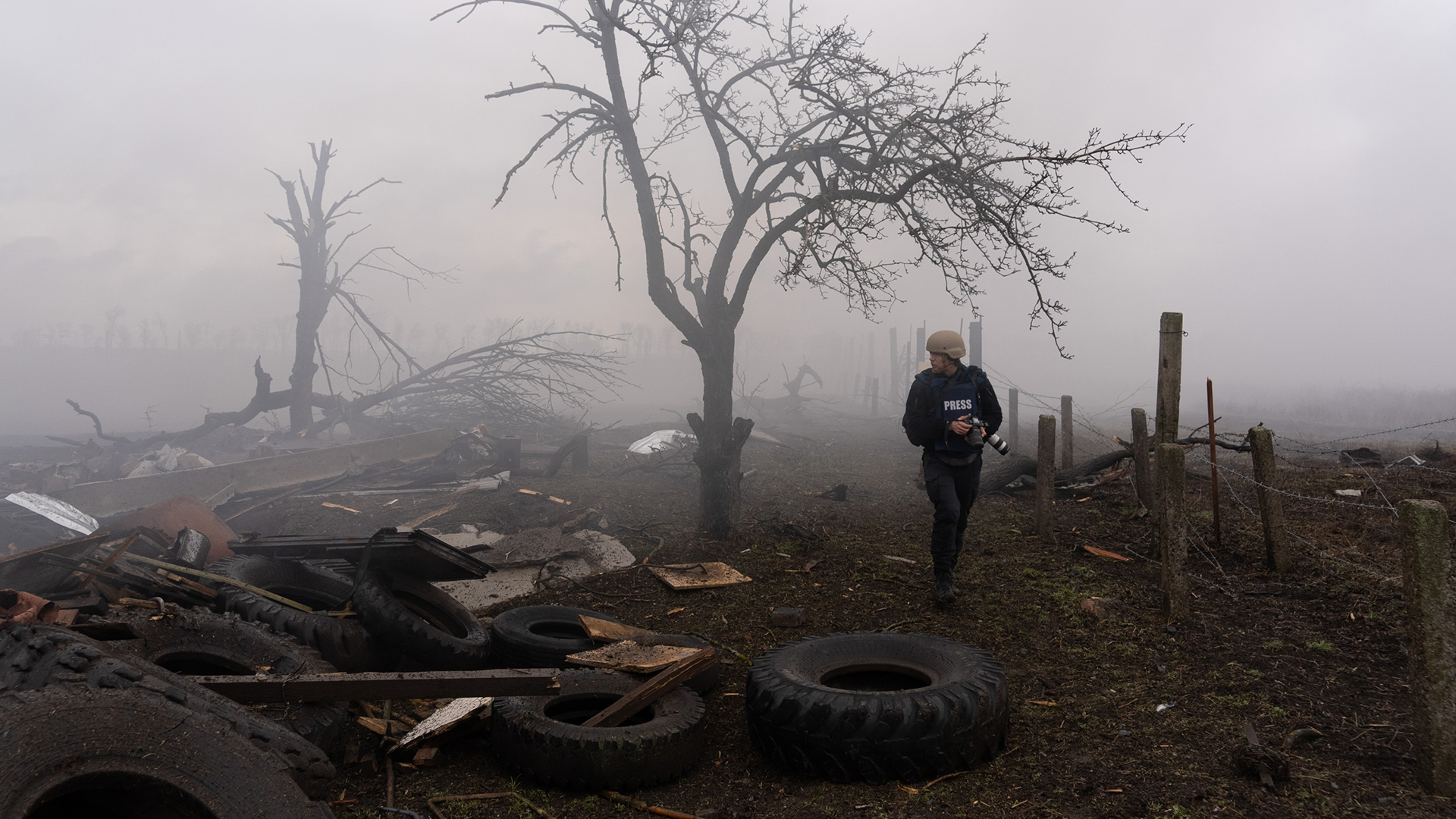 A journalist wearing a helmet and a jacket labeled 'PRESS' walking through a fog-covered, devastated landscape filled with discarded tires, broken wood, and debris. A bare, prominent tree stands amidst the mist, and remnants of a fence can be seen in the background.