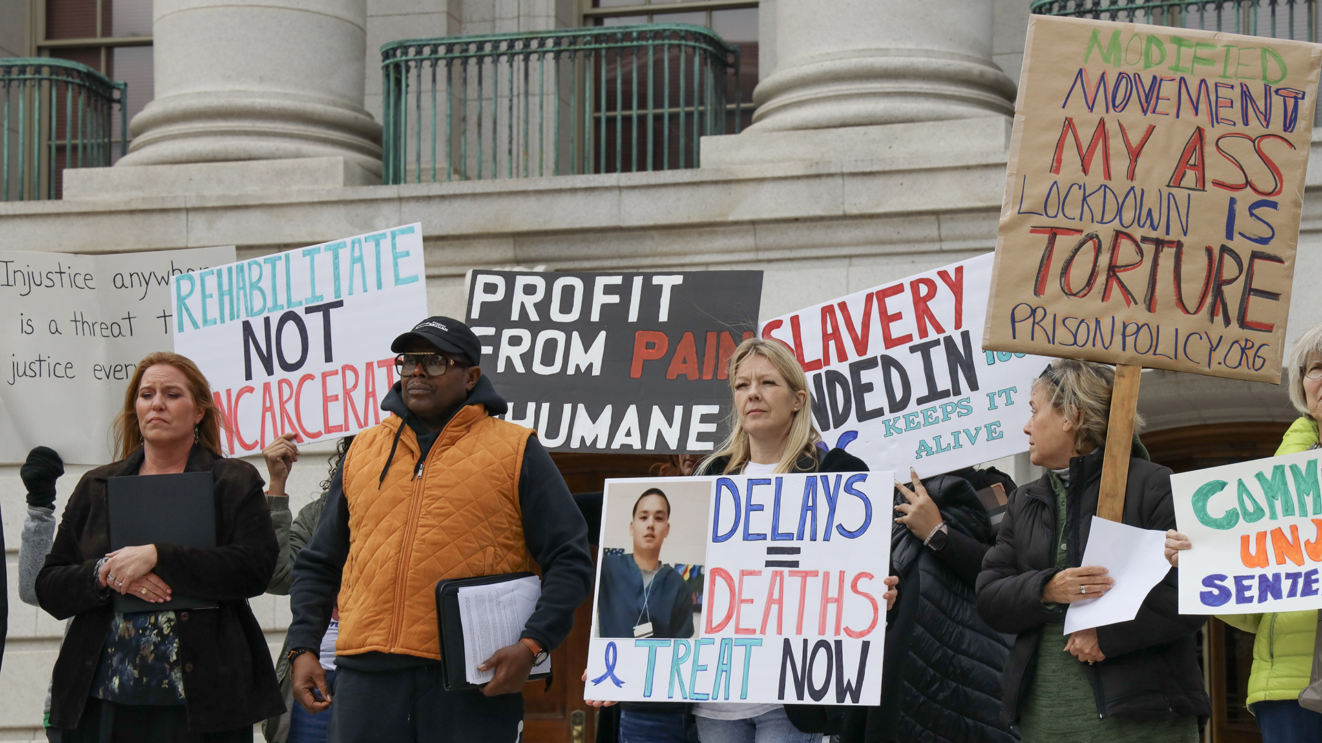 People stand in front of a building with marble masonry and pillars and hold signs that read "Rehabilitate Not Incarcerate," "Profit From Pain Inhumane," "Delays = Deaths Treat Now," and other phrases.