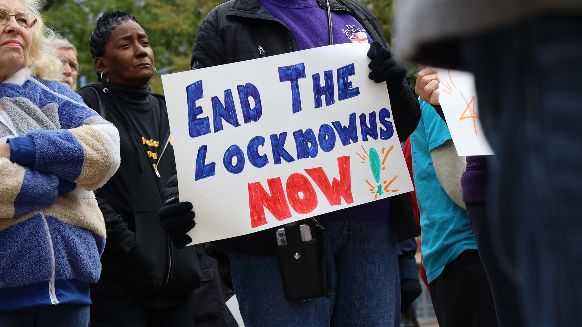 A person holds a hand-drawn sign reading "End the Lockdowns Now!" while standing among a group of people outside.
