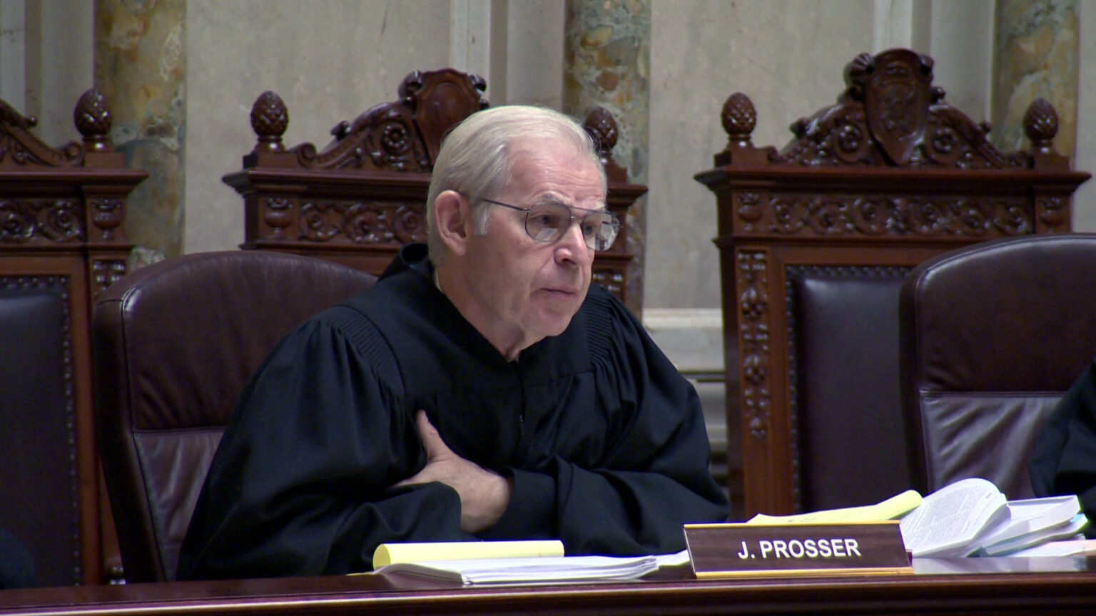 David Prosser sits in a high-backed leather chair with his arms folded on a judicial dais, with multiple papers and a nameplate reading J. Prosser on its surface, with multiple wood and leather high-backed chairs in the background, in a room with marble masonry.