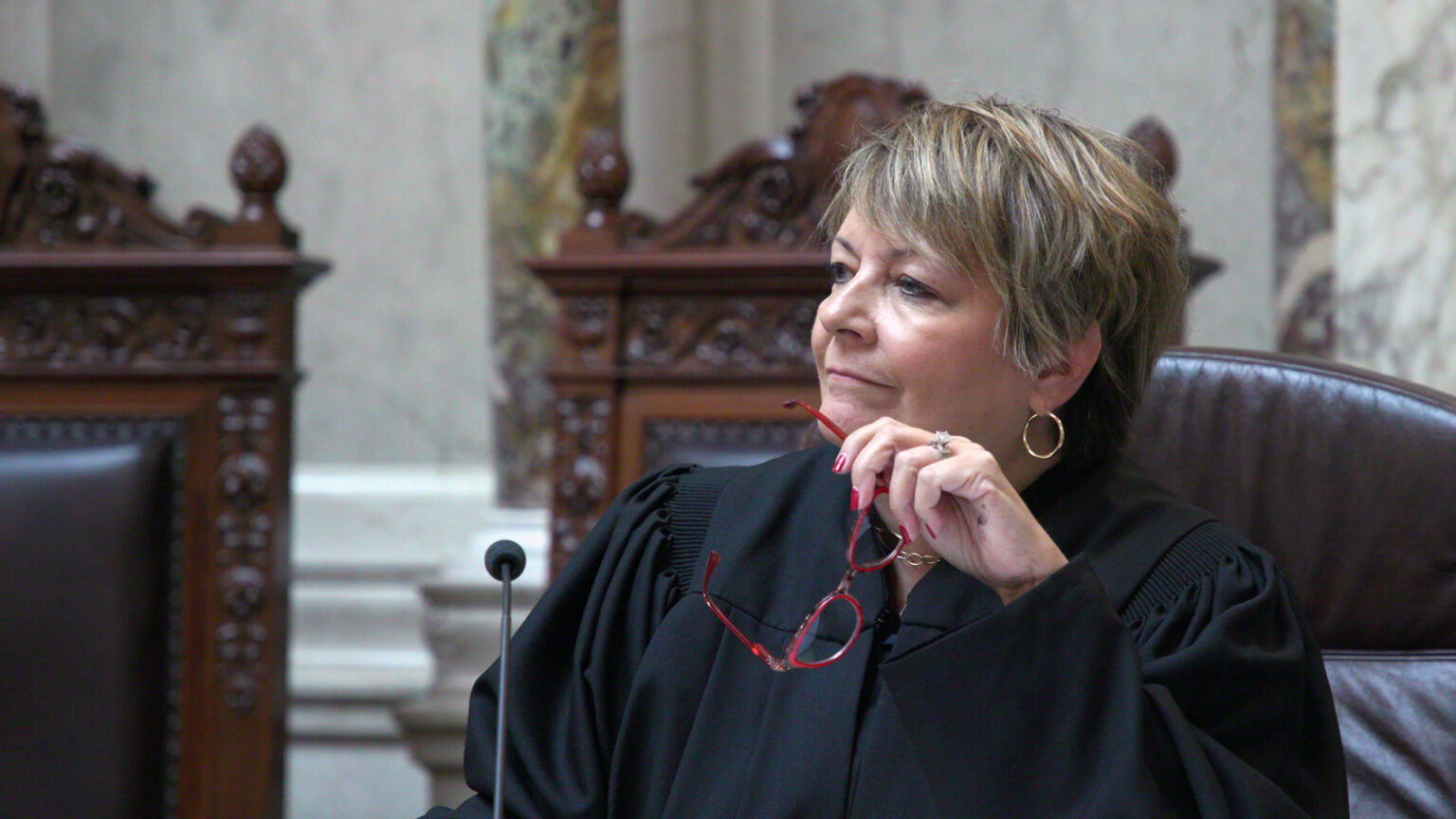 Janet Protasiewicz holds a pair of glasses in her left hand while sitting in a high-backed leather chair, with other high-backed wood and leather chairs in the background, in a room with marble masonry.