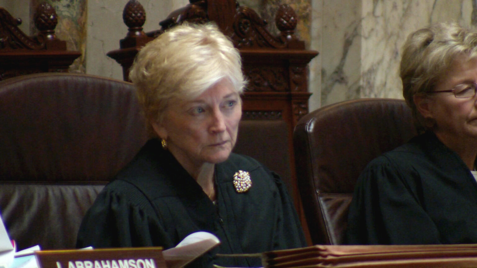 Patience Roggensack sits in a high-backed leather chair and listens, with another person seated to her side and empty high-backed wood and leather chairs behind her, in a room with marble masonry.