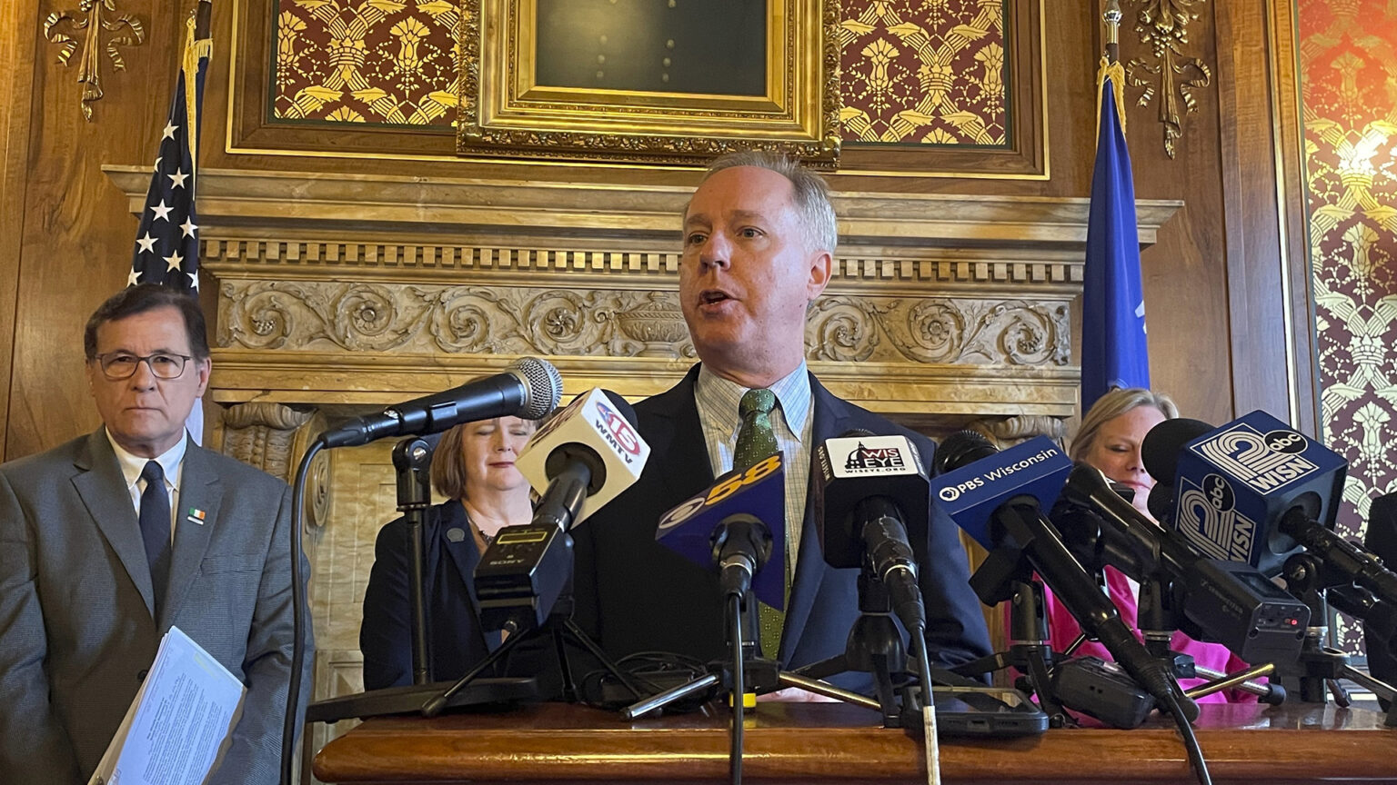 Robin Vos stands and speaks into multiple microphones with the flags of media organizations mounted to the top of a wood podium, with other people standing behind him, in a room with wood-paneled walls and toile wallpaper, a painting, and the U.S. and Wisconsin flags.