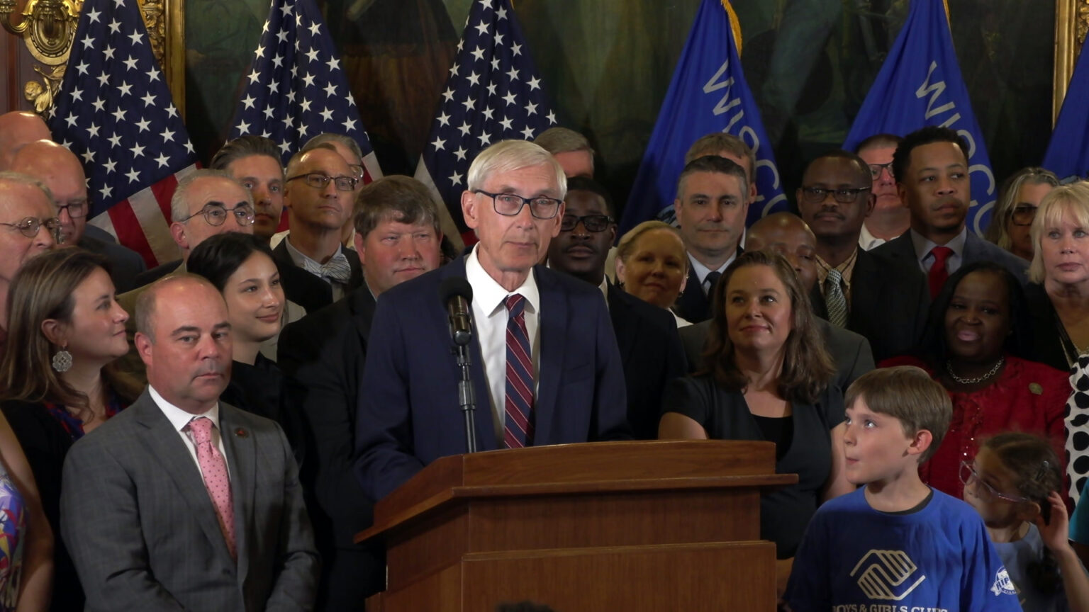 Tony Evers stands and speaks into a microphone mounted on a wood podium, with people standing behind him and to his side, and more people seated facing him, in a room with U.S. and Wisconsin flags in front of a large painting.