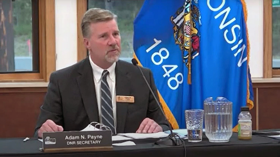 Adam Payne speaks into a microphone placed on a table with a nameplate reading Adam N. Payne and DNR Secretary, a glass and pitcher of water and other items, with a Wisconsin flag in the background and windows facing a wooded area.