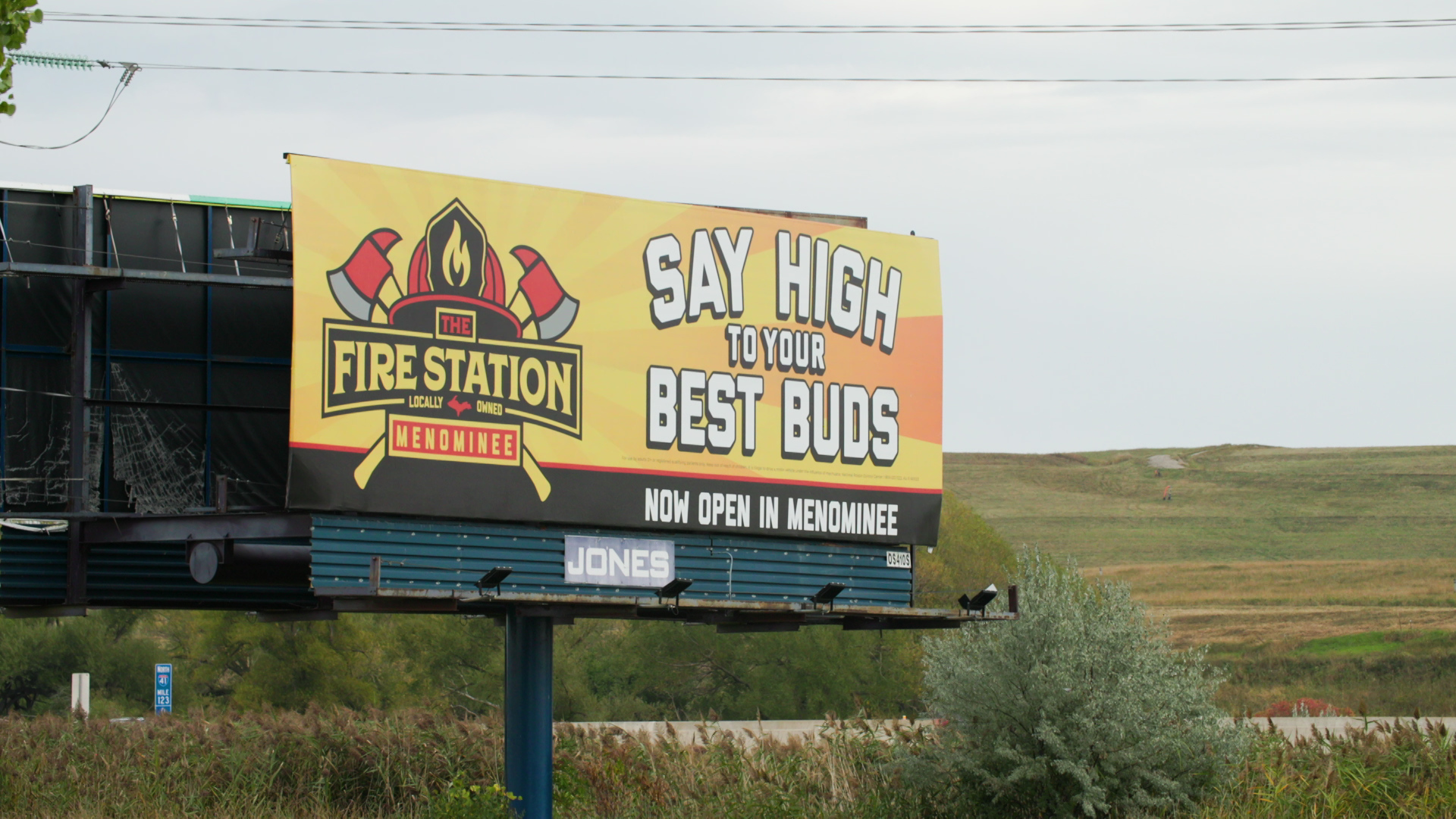 A billboard with the words "Say High to Your Best Buds" and "Now Open in Menominee" and a "The Fire Station" marijuana dispensary logo with a helmet and crossed axes stands next to a highway, with grass-covered hillsides in the background.