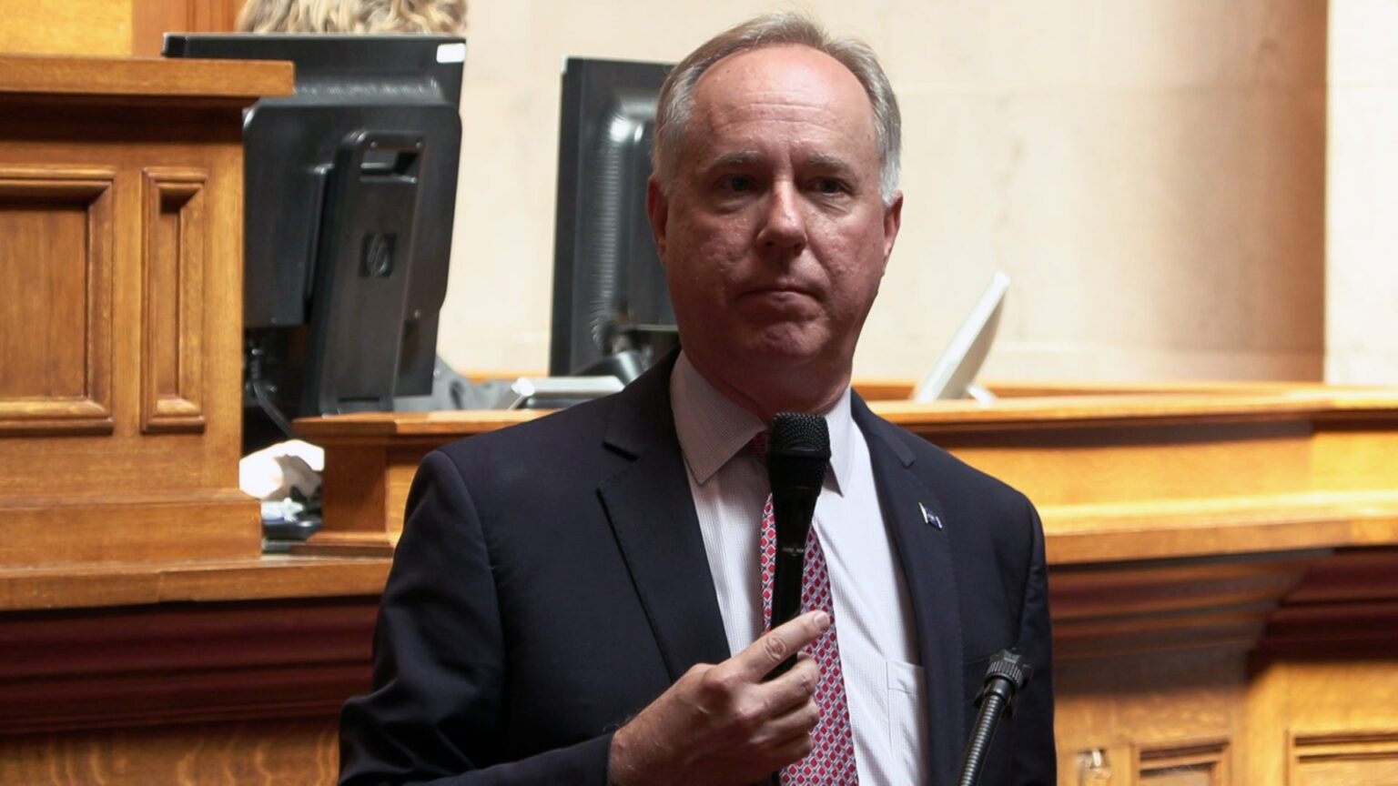 Robin Vos speaks into a microphone while standing in front of a wood legislative dais, with the back of two computer monitors on its surface in the background.