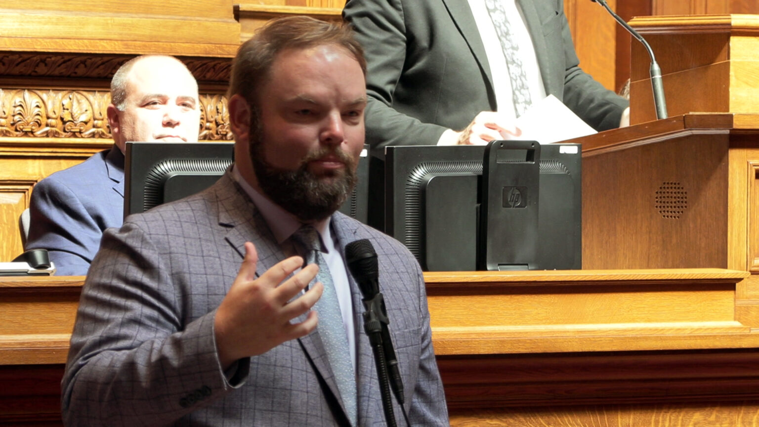 Tyler August gestures with his right hand while speaking in a microphone, with one person seated and another standing on a wood judicial dais in the background.