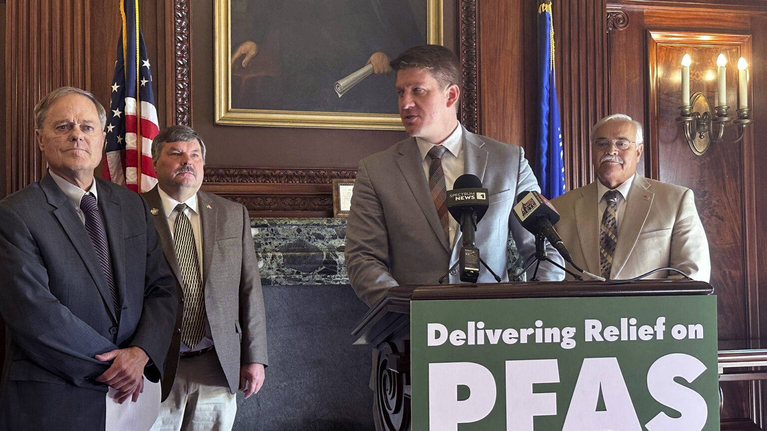Eric Wimberger looks to the right while standing behind a podium affixed with two microphones and a sign on its front reading Delivering Relief on PFAS, with Rob Cowles and Rob Swearingen on one side and Jeff Mursau on the other, in a room with wood-paneled walls, the U.S. and Wisconsin flags, an painting, and an illuminated electric wall sconce.