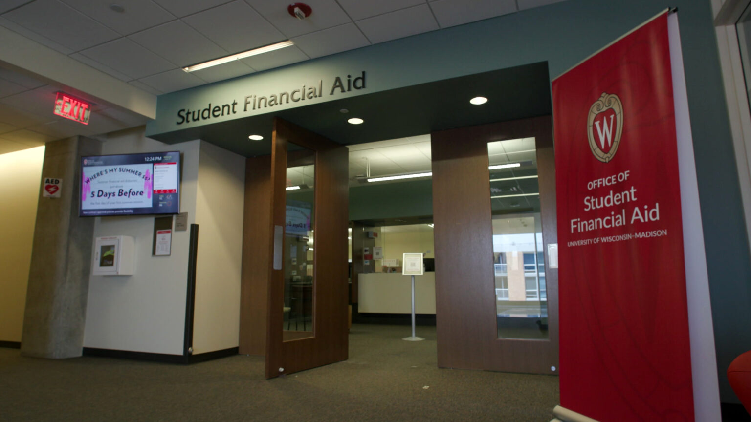 A vinyl banner with words Office of Student Financial Aid and University of Wisconsin-Madison with the university's 'W' crest on it stands in a hallway, next to glass-paneled door under a word sign reading Student Financial Aid.