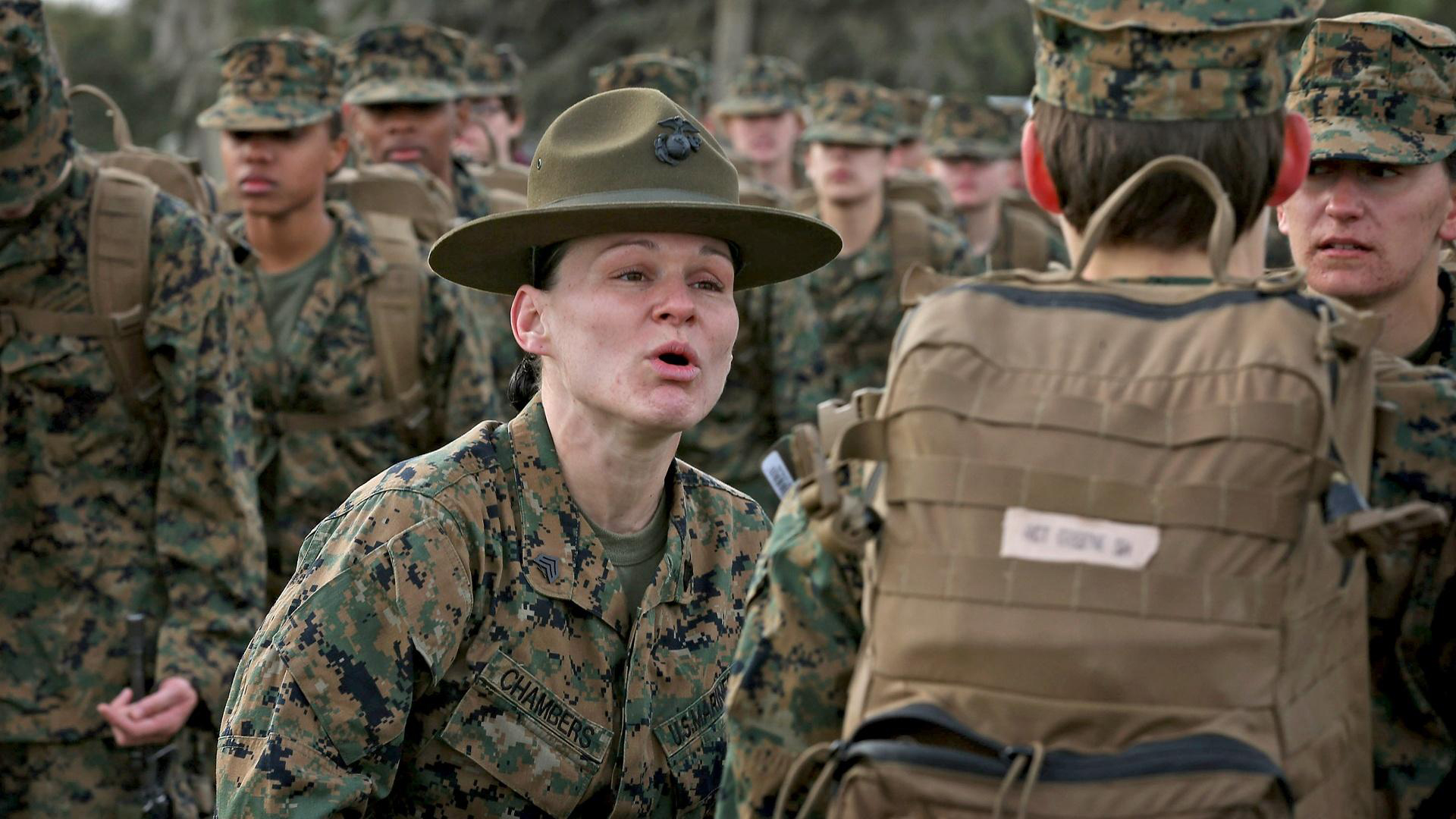 A female Marine in a brown hat, identified by the name "Chambers" on her uniform, speaks to a group of Marines in camouflage. They stand attentively, some with backpacks, in an outdoor setting.