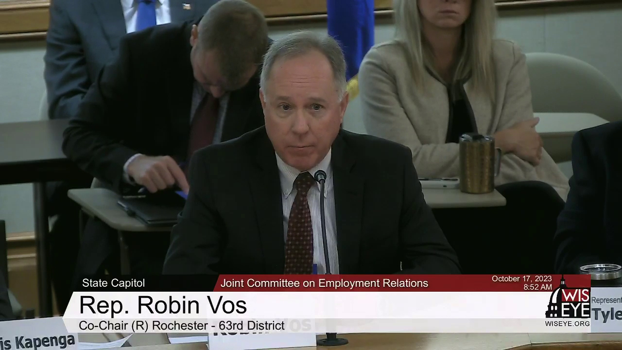 A video still image shows Robin Vois sitting at a table while speaking into a microphone in a room with other people seated in the background, with a video graphic at bottom including the text Rep. Robin Vos, Co-Chair (R) Rochester - 63rd District and Joint Committee on Employment Relations.