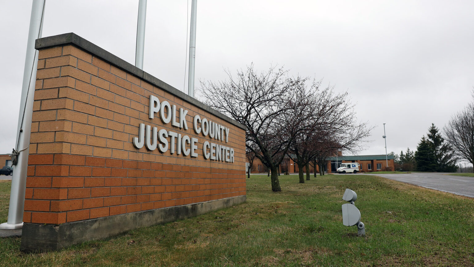 A short brick wall with a letter sign reading Polk County Justice Center stands on a lawn next to three flagpoles and in front of a row of leafless trees leading to a building with a U.S. Postal Service delivery truck parked in front, with more trees in the background.