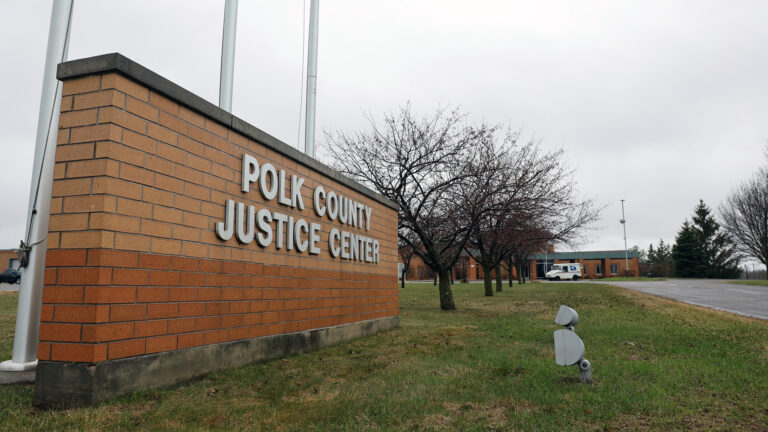 A short brick wall with a letter sign reading Polk County Justice Center stands on a lawn next to three flagpoles and in front of a row of leafless trees leading to a building with a U.S. Postal Service delivery truck parked in front, with more trees in the background.