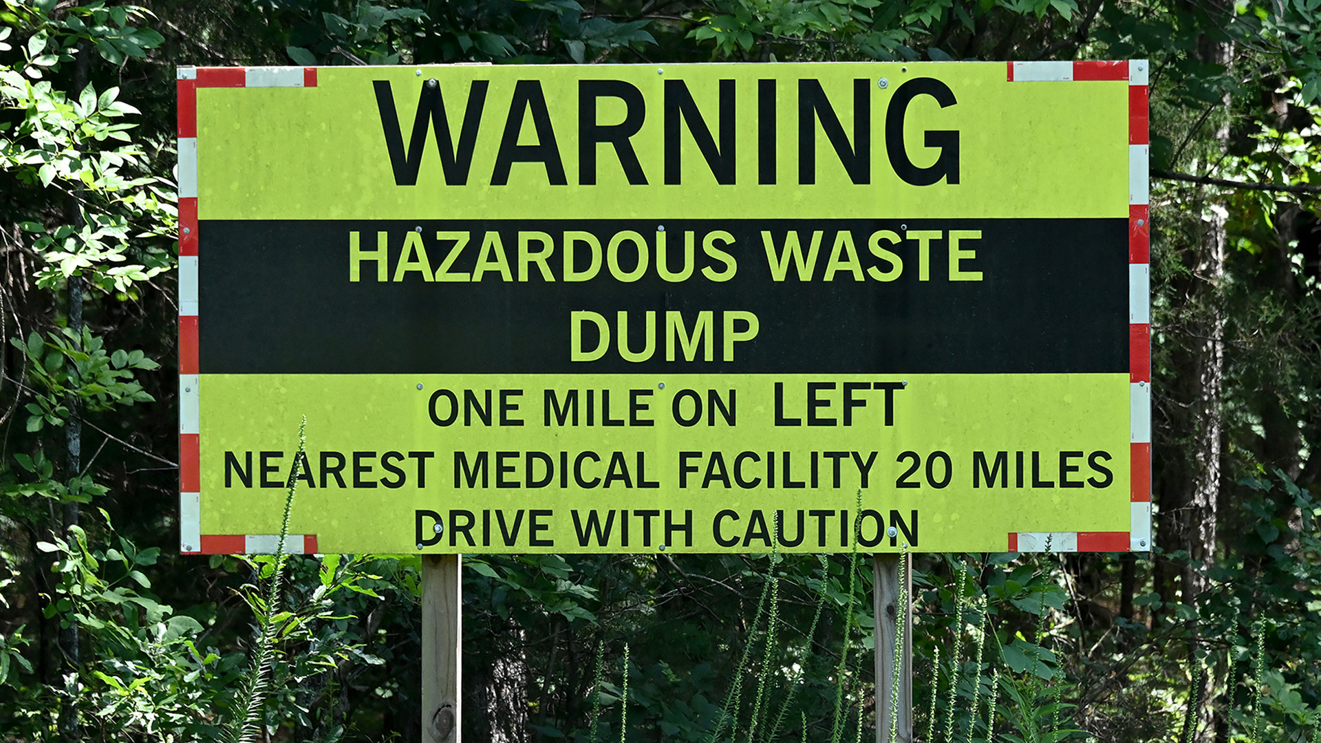 A sign with the words "Warning," "Hazardous Waste Dump," One Mile on Left," "Nearest Medical Facility 20 Miles," and "Drive With Caution" stands in front of trees and ground foliage.