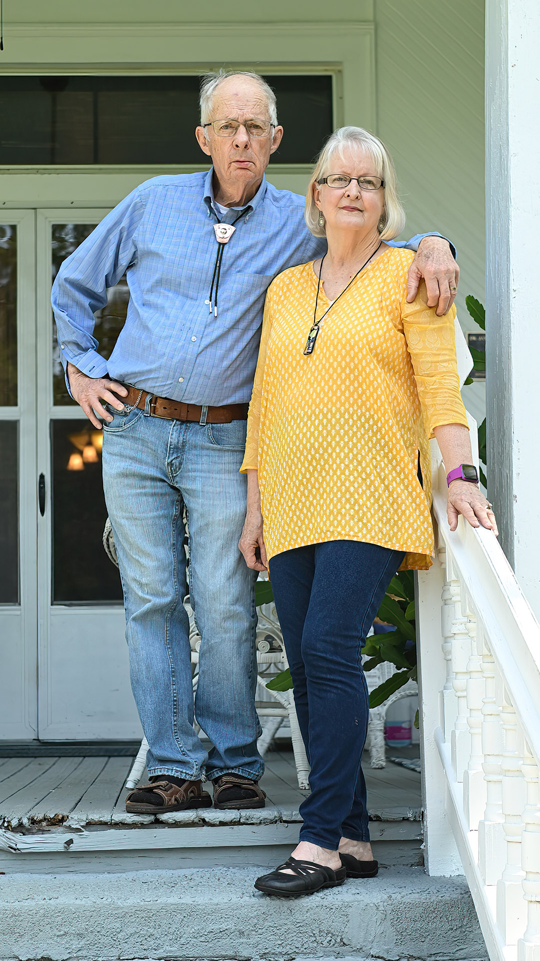 Linda Munoz and Charles Munoz pose for a portrait while standing next to a railing on a porch in front of a double-door entrance to a building.