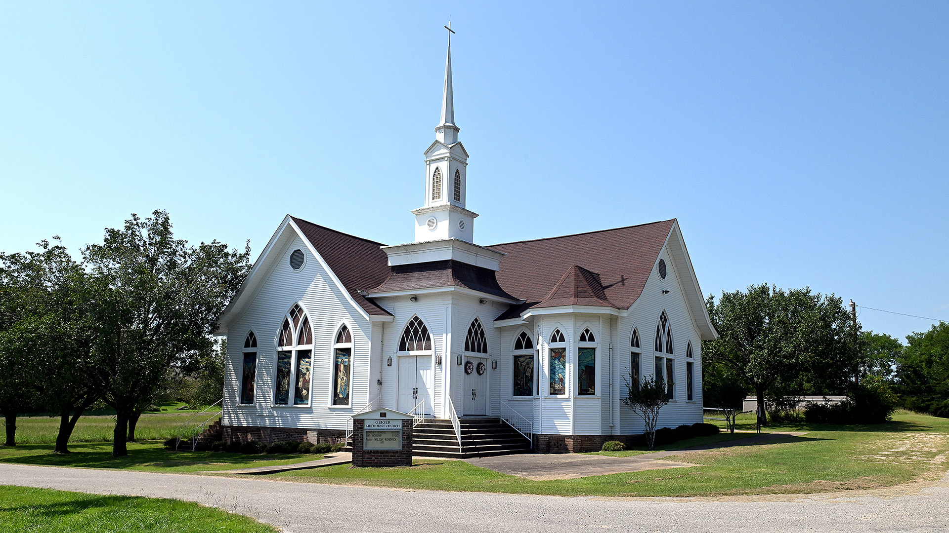 A wood-sided church with a steeple topped by a cross and flanked by two building wings with stained-glass windows stands next to a driveway and is surrounded by trees.