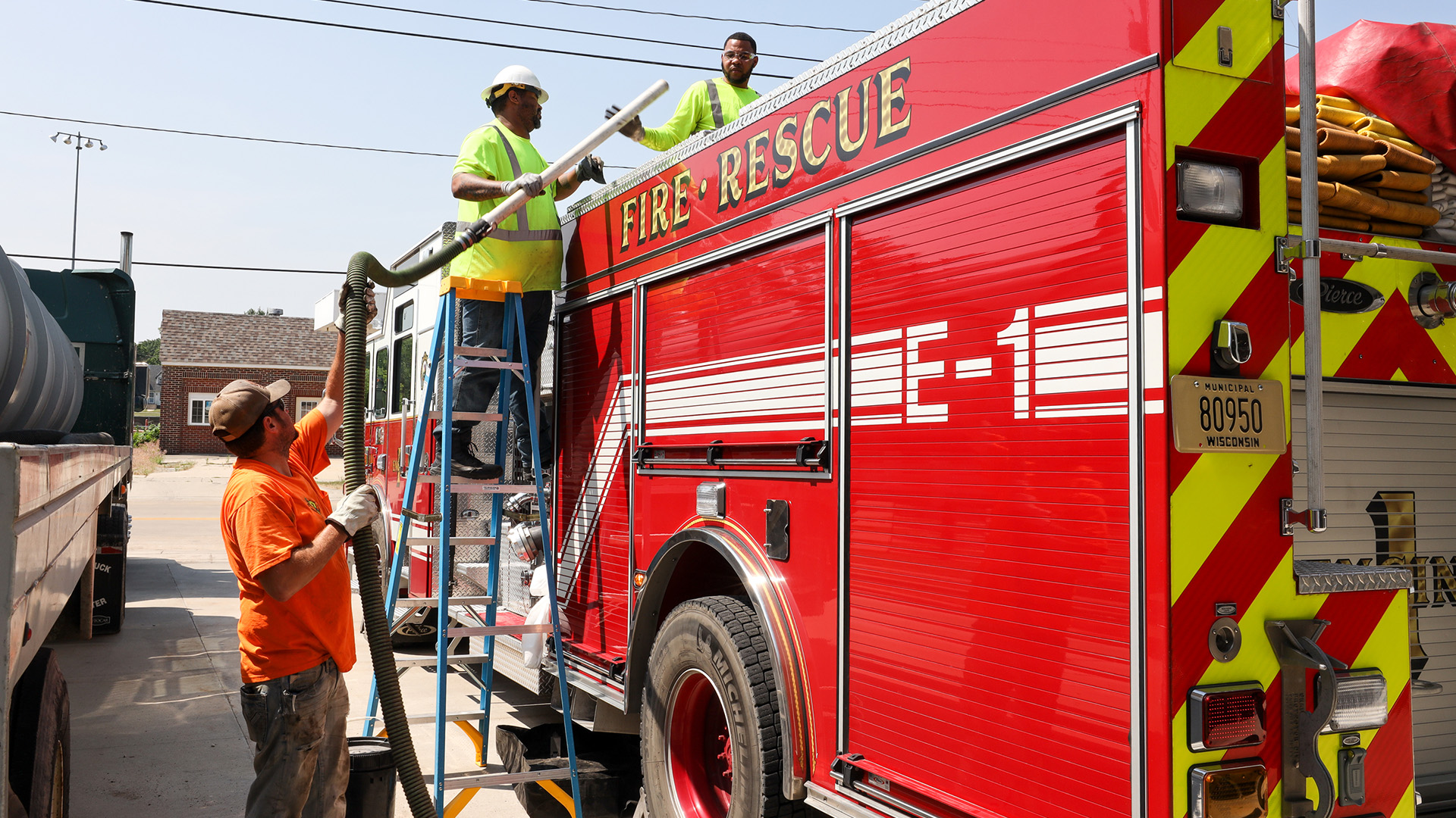 Dan Bice holds a flexible corrugated plastic hose up to Tyrone Rogers, who is standing on a folding ladder placed next to a fire and rescue truck, with Trent Thomas on its top holding its nozzle, with another truck parked to the side and a building and power lines in the background.