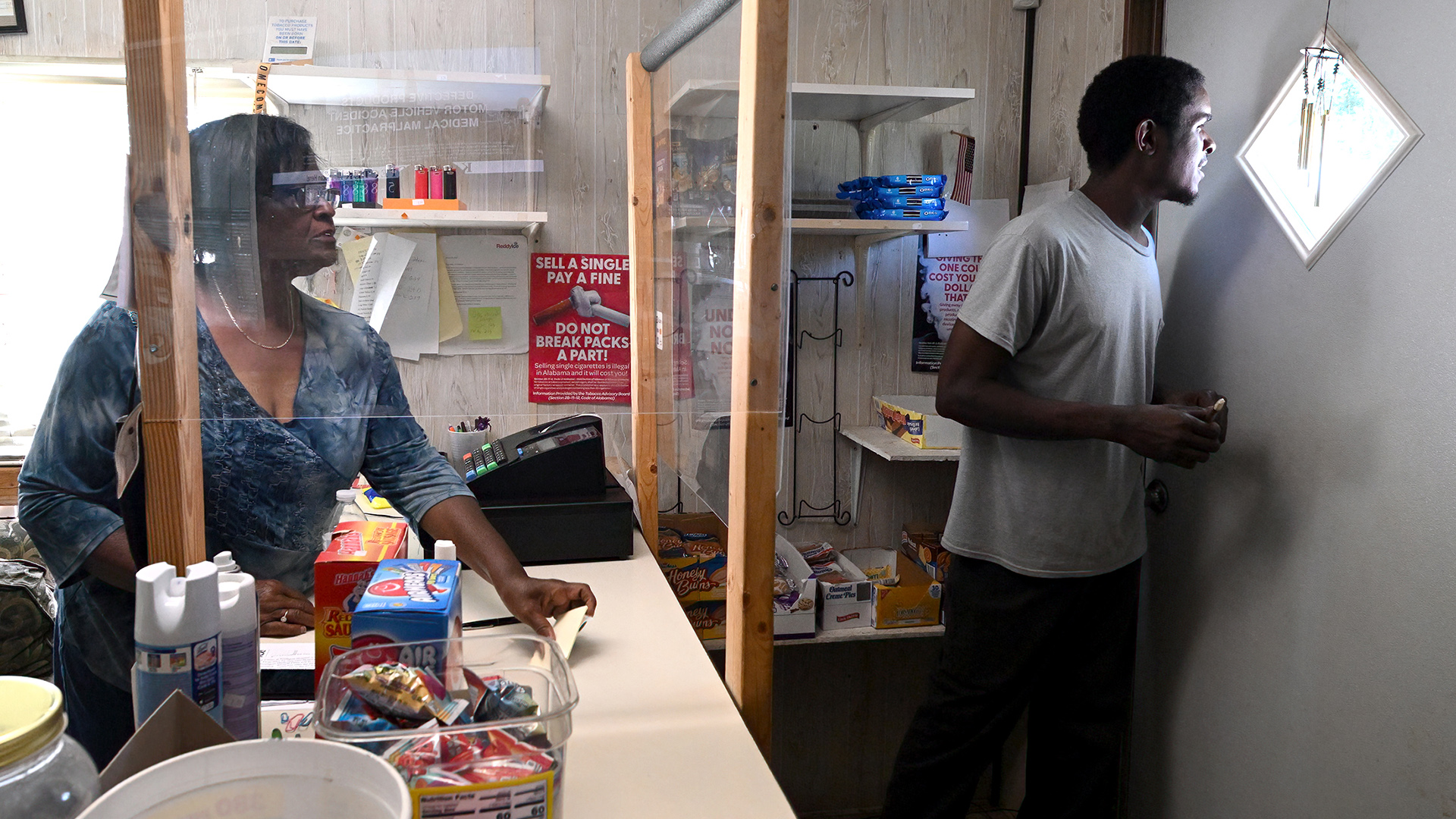 Dorothy Oliver stands behind a counter with a cash register and multiple items for sale, with a wood-and-transparent plastic sanitary barrier mounted on two sides, while Cedric Magee looks out a square window at a 45-degree angle in the top of a door.